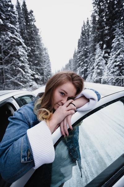 冬天，一个穿着蓝色牛仔服的女人靠在路上白雪皑皑的松树旁的车门上